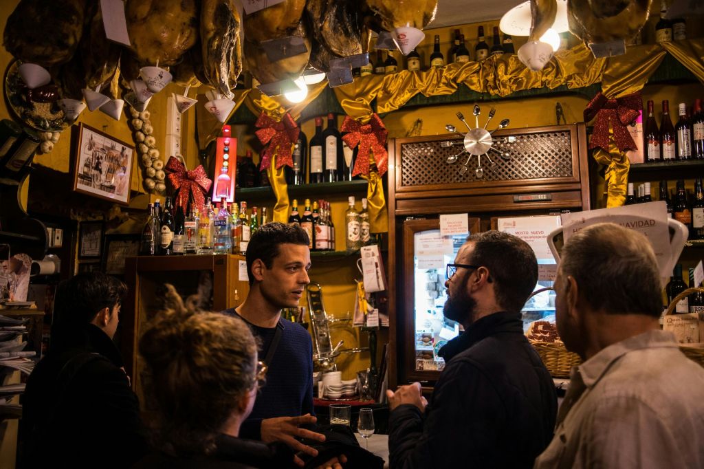 경화 고기, 붉은 활, 술과 와인 병이 놓인 선반이 놓인 바에서 네 명이 이야기를 나누고 있습니다.