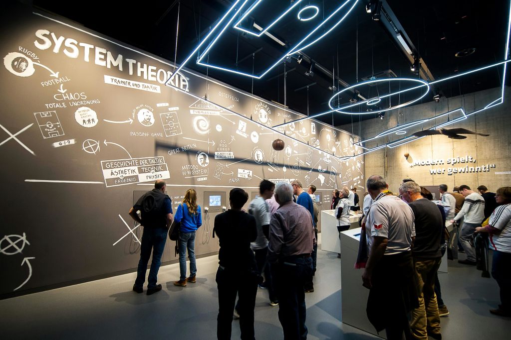 현대적인 전시 공간에서 다이어그램과 텍스트가 있는 벽을 보는 사람들은 천장에 설치된 네온 블루 조명으로 조명을 받습니다.