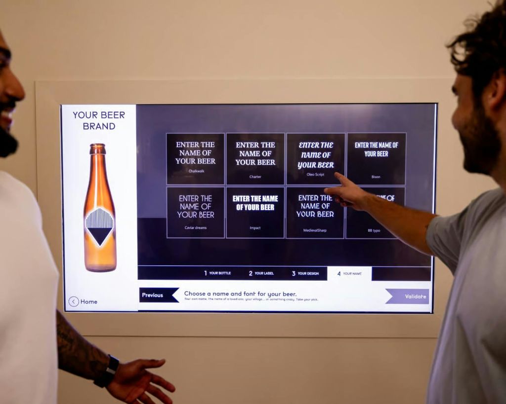 맥주 이름을 입력하고 디자인을 선택할 수 있는 옵션을 포함하여 맥주 사용자 정의 인터페이스를 표시하는 터치스크린과 상호 작용하는 두 사람.