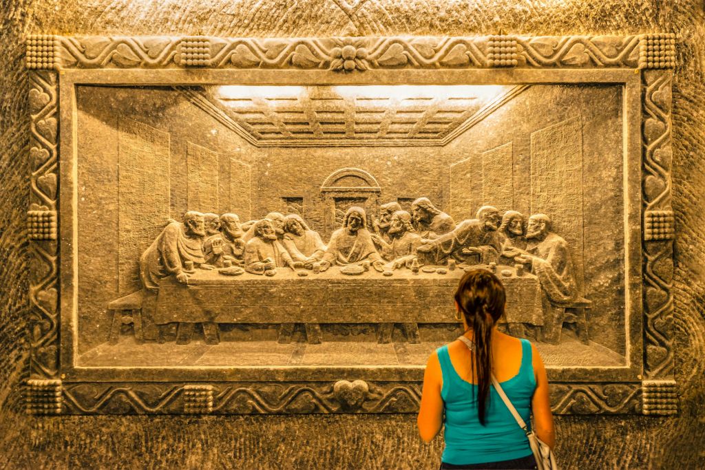 청록색 상의를 입은 한 여성이 벽에 '최후의 만찬' 을 세밀하게 조각한 돌을 바라보고 있습니다.