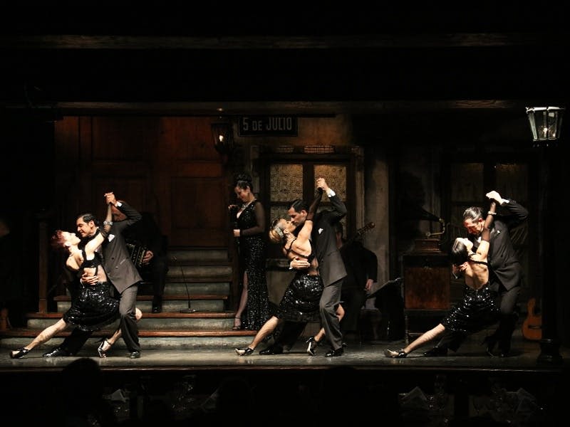나무 문, 계단, '5 de Julio' 표지판을 배경으로 조명이 어두운 무대에서 검은색 복장을 한 커플이 탱고를 연주합니다.