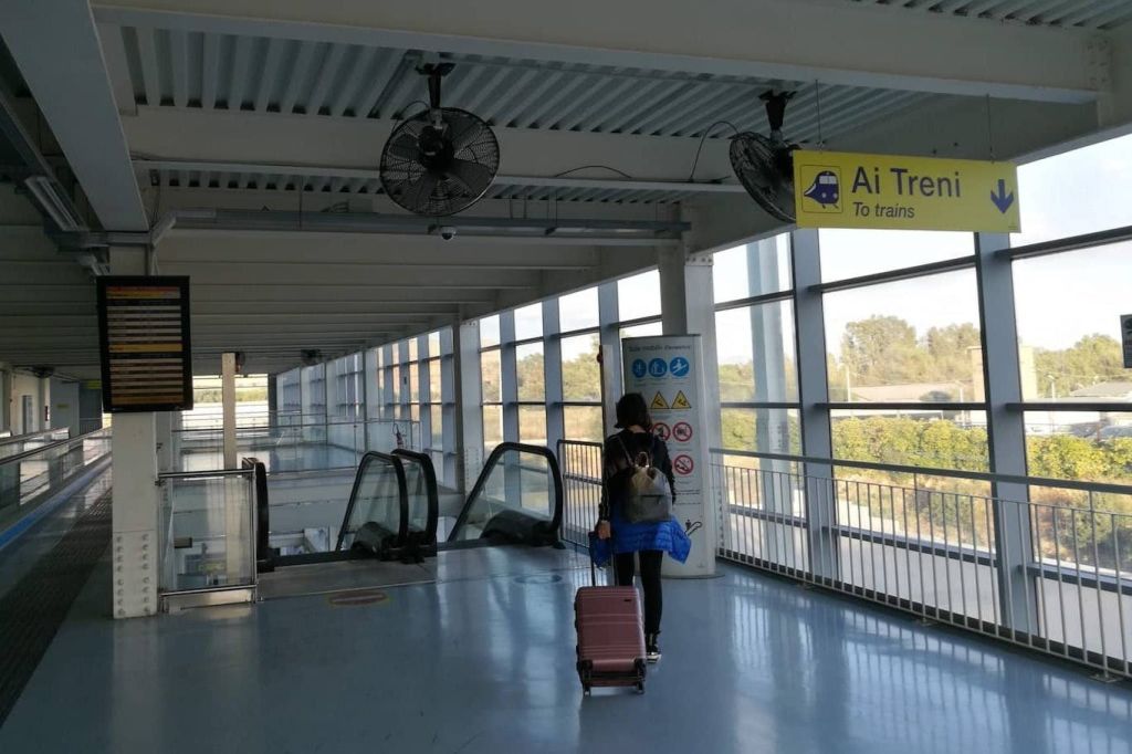 현대적인 교통 터미널에서 에스컬레이터를 향해 걸어가는 짐을 가진 사람. 표지판에는 열차를 가리키는 "Ai Treni" 라고 쓰여 있습니다.
