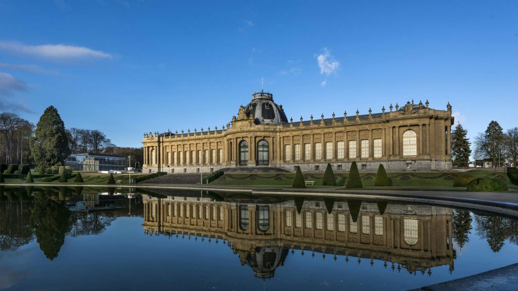 돔, 커다란 창문, 잘 다듬어진 관목이 있는 화려한 역사적인 건물로, 맑고 푸른 하늘 아래 잔잔한 연못에 반사됩니다.