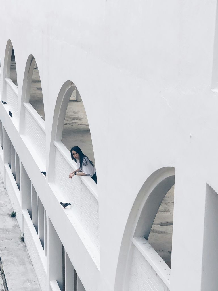 커다란 아치가 있는 하얀 건물의 난간에 사람이 기대고 있습니다. 새들은 아치의 선반에 자리 잡고 있습니다.