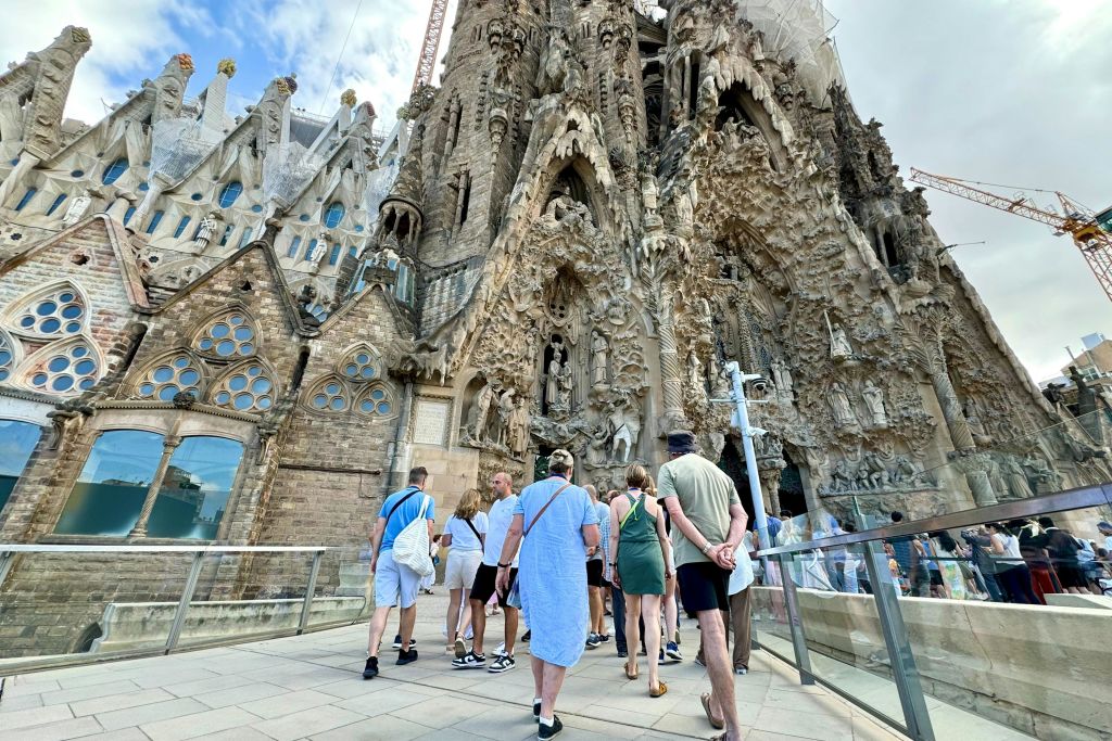 관광객들은 화창한 날 라 사그라다 파밀리아 대성당의 정교하게 조각된 정면을 향해 걸어가고 있습니다.