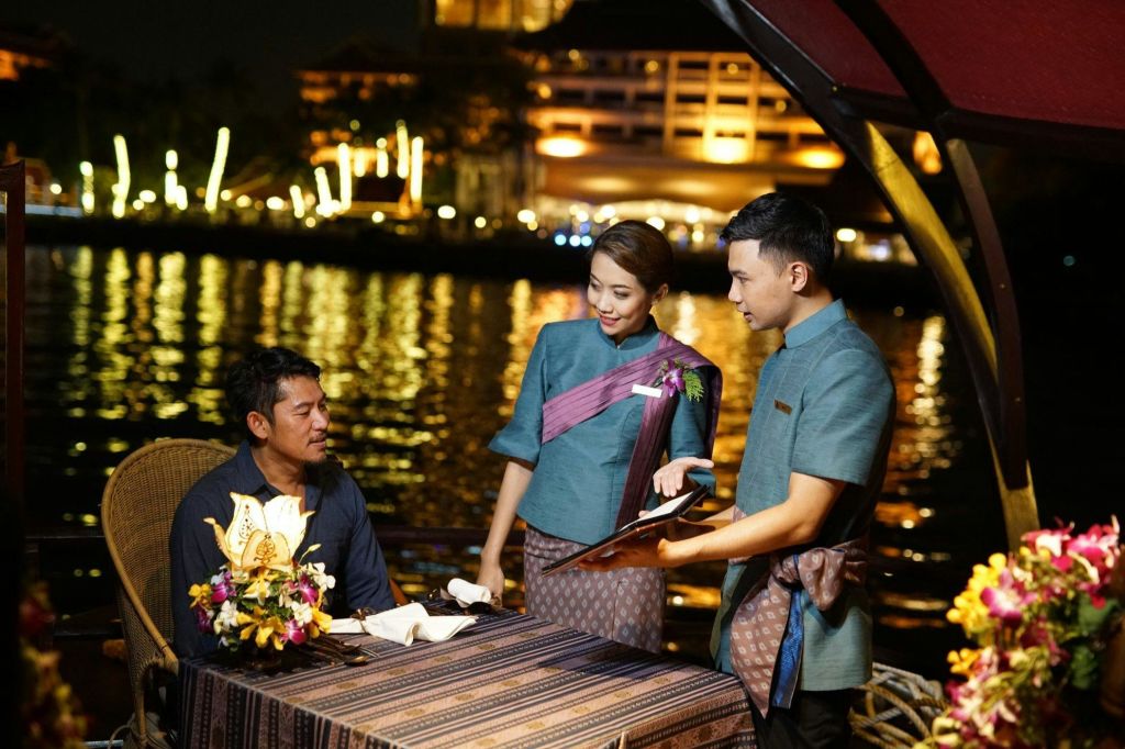 한 남성이 강변 레스토랑에서 저녁 조명 아래에서 전통 복장을 한 두 명의 직원과 대화를 나누고 있습니다.