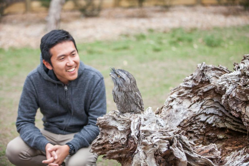 한 사람이 웅크리고 앉아 야외 공간의 나무 조각 위에 앉아 있는 새를 바라보며 미소를 짓고 있습니다.