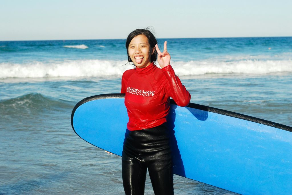 얕은 바닷물에 서서 파란색 서핑 보드를 들고 빨간색 "Ride-A-Wave" 셔츠를 입고 손짓을 하는 사람.