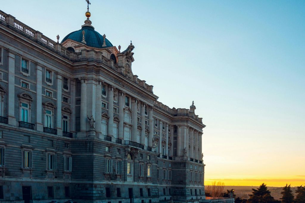 석양의 하늘을 배경으로 화려한 건축물과 푸른 돔이 있는 역사적인 대형 건물입니다.