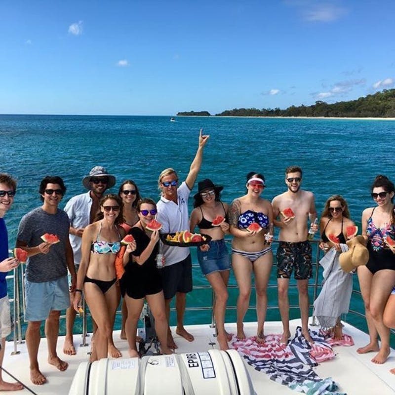 화창한 날 보트에서 수박 조각을 들고 있는 수영복을 입은 한 무리의 사람들. 바다와 먼 해안선을 배경으로 말이죠.