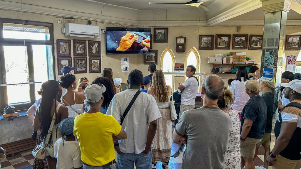 아치형 창문이 있고 선반에 다양한 물품이 놓여 있는 방에서 TV로 교육용 비디오를 시청하는 사람들의 모임.