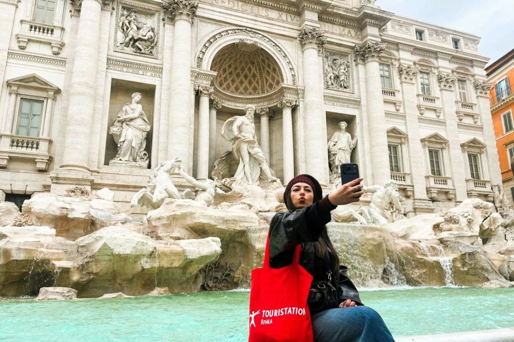 한 사람이 로마의 트레비 분수 앞에서 "TOURISTATION ROME" 이 인쇄된 빨간 가방을 들고 셀카를 찍고 있습니다.