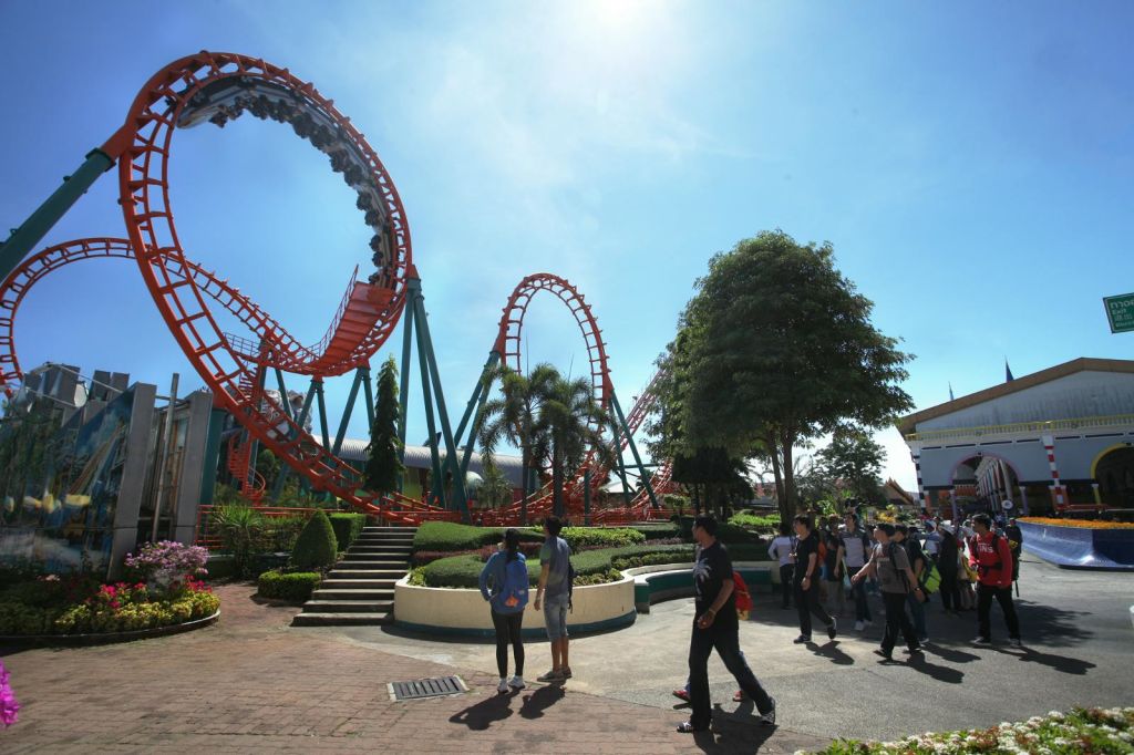 화창한 날 공원의 빨간 롤러코스터 근처를 걷는 사람들. 나무와 조경된 녹지가 있습니다.