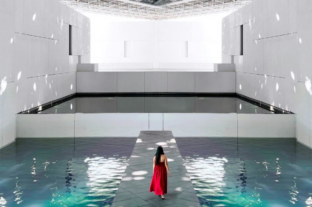 붉은 드레스를 입은 한 여성이 물로 둘러싸인 좁은 길을 걸으며 현대적이고 하얀 건축 공간에 들어섭니다.