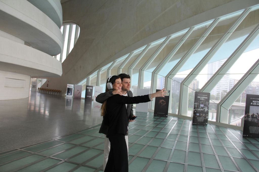 커다란 창문이 있는 현대적인 건물 내부에서 두 사람이 셀카를 찍고 있습니다. 하나는 헤드폰을 착용하고 있습니다. 디스플레이 패널이 배경에 있습니다.