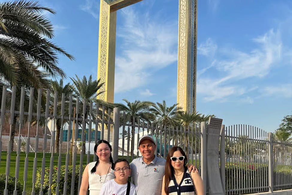 야자수가 있는 화창한 푸른 하늘 아래 직사각형의 커다란 황금 프레임 구조물 앞에서 포즈를 취하고 있는 네 사람.