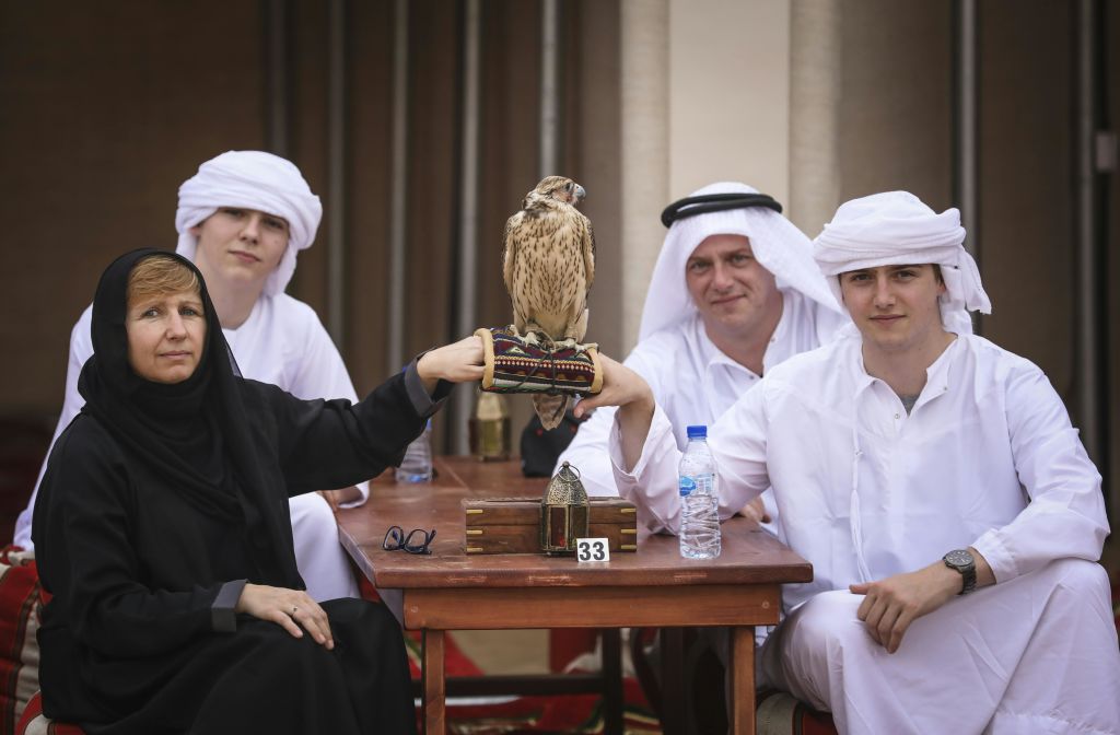 세 사람이 테이블에 앉아 장갑을 낀 여성의 손에 매를 얹고 있습니다. 테이블에는 물병과 안경이 있습니다.