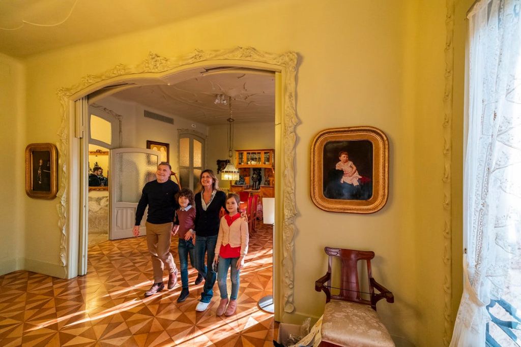 화려한 장식, 나무 바닥, 벽에 액자 그림이 있는 따뜻한 조명으로 꾸며진 방에는 4인 가족이 서 있습니다.