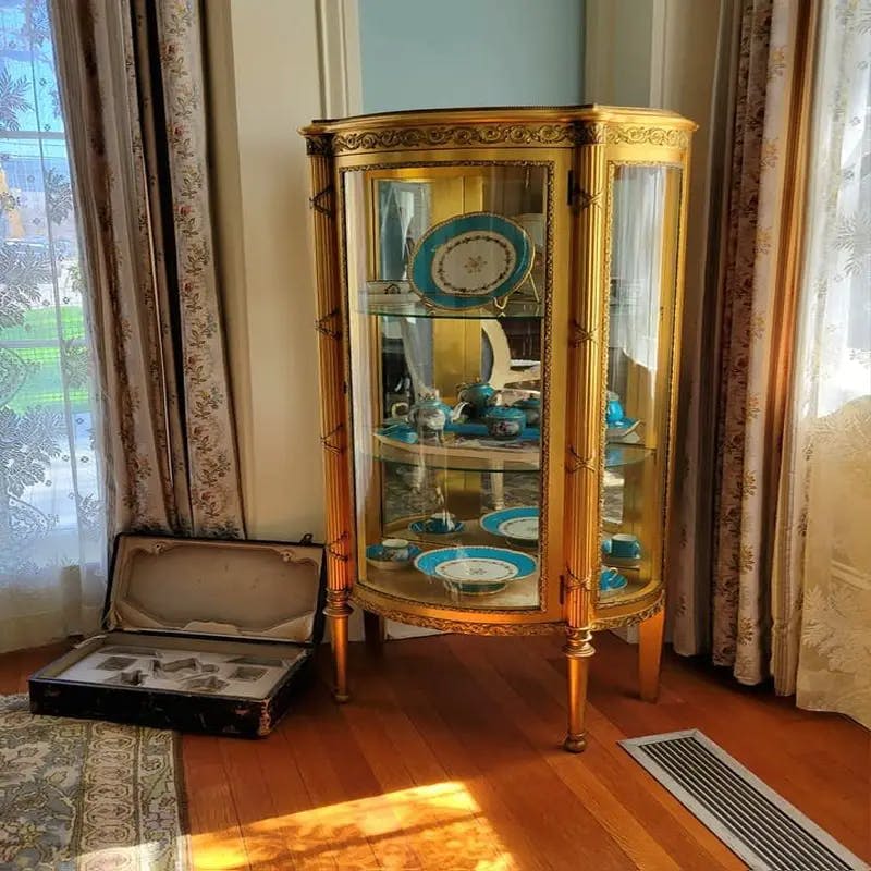 나무 바닥에 열린 여행 가방 옆에 다양한 파란색과 흰색 식기를 수납할 수 있는 금장 장식이 있는 유리 디스플레이 캐비닛.