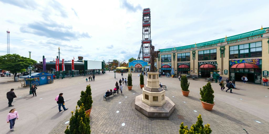 중앙에 동상, 화분, 벤치, 건물이 있는 공공 광장. 높은 놀이공원을 배경으로 즐길 거리가 마련되어 있습니다.