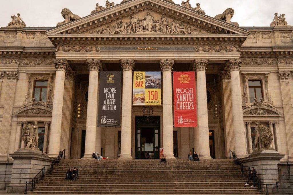 맥주 박물관을 광고하는 기둥과 배너가 있는 고전적인 건물로 앞 계단에 사람들이 앉아 있습니다.