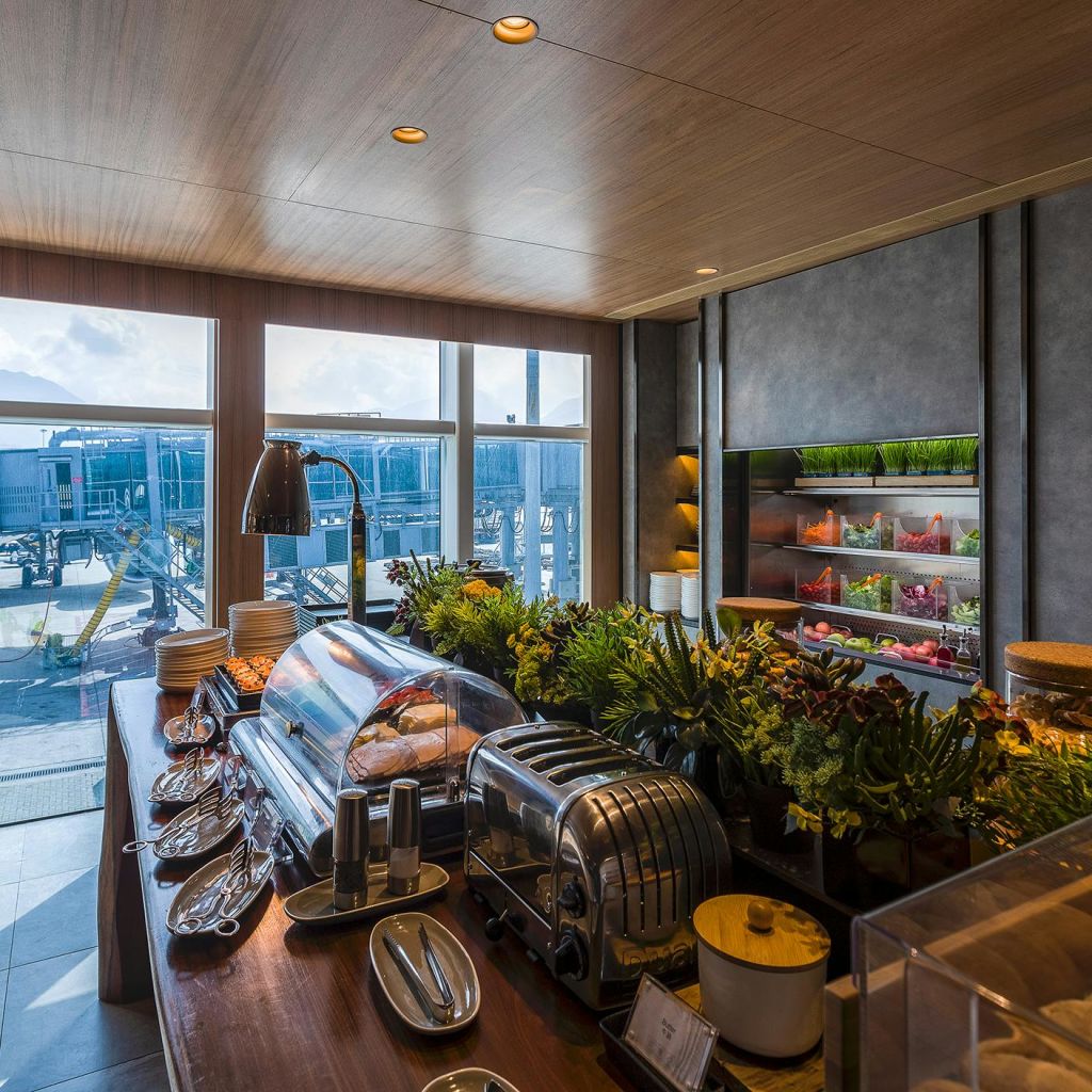 공항 활주로가 내려다보이는 커다란 창문 근처에 덮개 덮인 요리, 반짝이는 토스터, 신선한 야채가 있는 뷔페 세트가 있습니다.