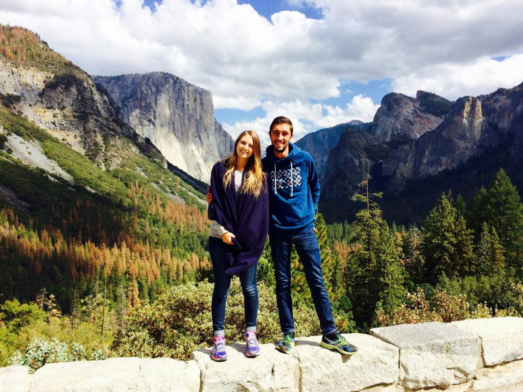 아름다운 산 풍경과 흐린 하늘을 배경으로 석조 선반에 미소 짓는 커플이 서 있습니다.