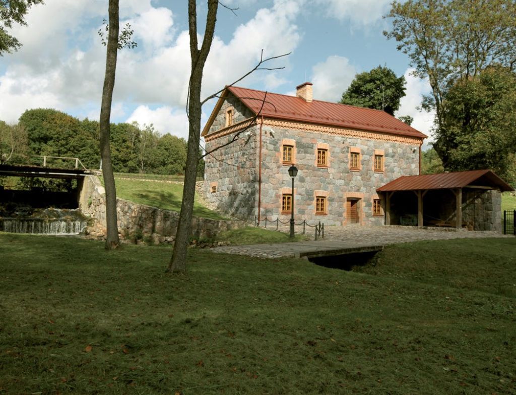 나무로 둘러싸인 붉은 지붕, 잔디밭, 자갈길, 개울 위의 작은 다리가 있는 석조 주택.