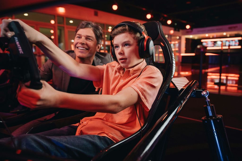 헤드폰을 착용한 주황색 셔츠를 입은 한 소년이 시뮬레이터 게임에 집중하고 있으며, 옆에서 한 노인이 미소를 짓고 있습니다.