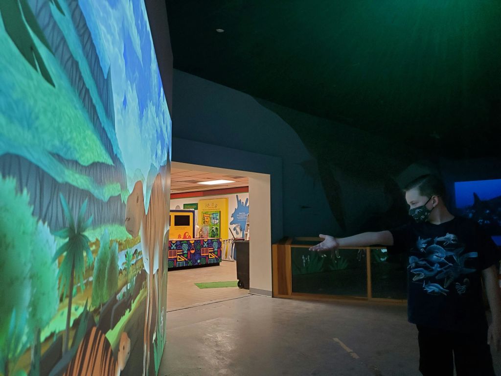 마스크를 쓴 어린이가 건물 내부의 선사 시대 풍경을 형형색색의 커다란 벽에 투영하여 상호 작용합니다.