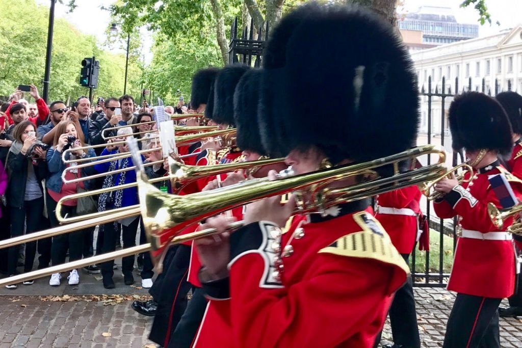 빨간색 유니폼과 곰 가죽 모자를 쓴 뮤지션이 황동 악기를 연주합니다. 많은 구경꾼이 장벽 뒤에서 사진을 찍습니다.
