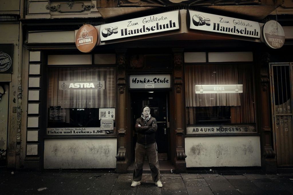 한 사람이 "Zum Goldenen Handschuh" 라는 이름의 조명이 희미한 바 앞에 팔짱을 끼고 서 있습니다. 창문에 간판과 커튼이 있습니다.