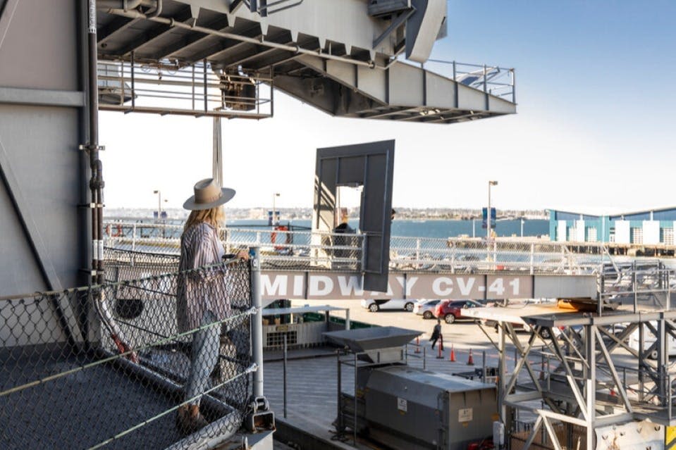 모자를 쓴 사람이 USS 미드웨이 CV-41에 탑승한 전망대에 서 있으며, 해안 풍경과 항구를 배경으로 합니다.