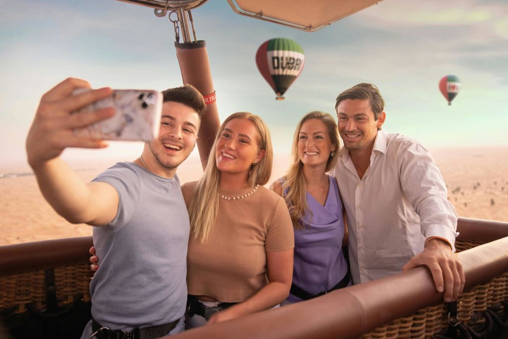 사막 풍경을 배경으로 다른 두 개의 열기구가 있는 열기구 바구니에서 네 명이 미소를 짓고 있습니다.