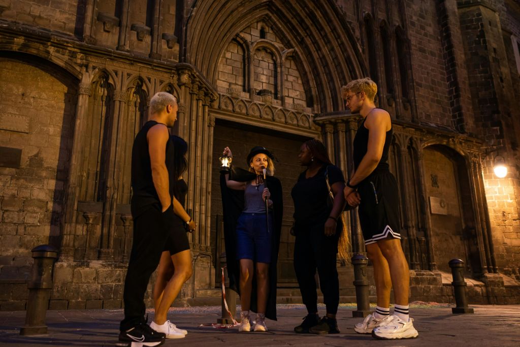 검은 망토를 입은 투어 가이드가 밤에 화려하고 커다란 석조 건물 밖에서 네 사람에게 말을 건넨다.