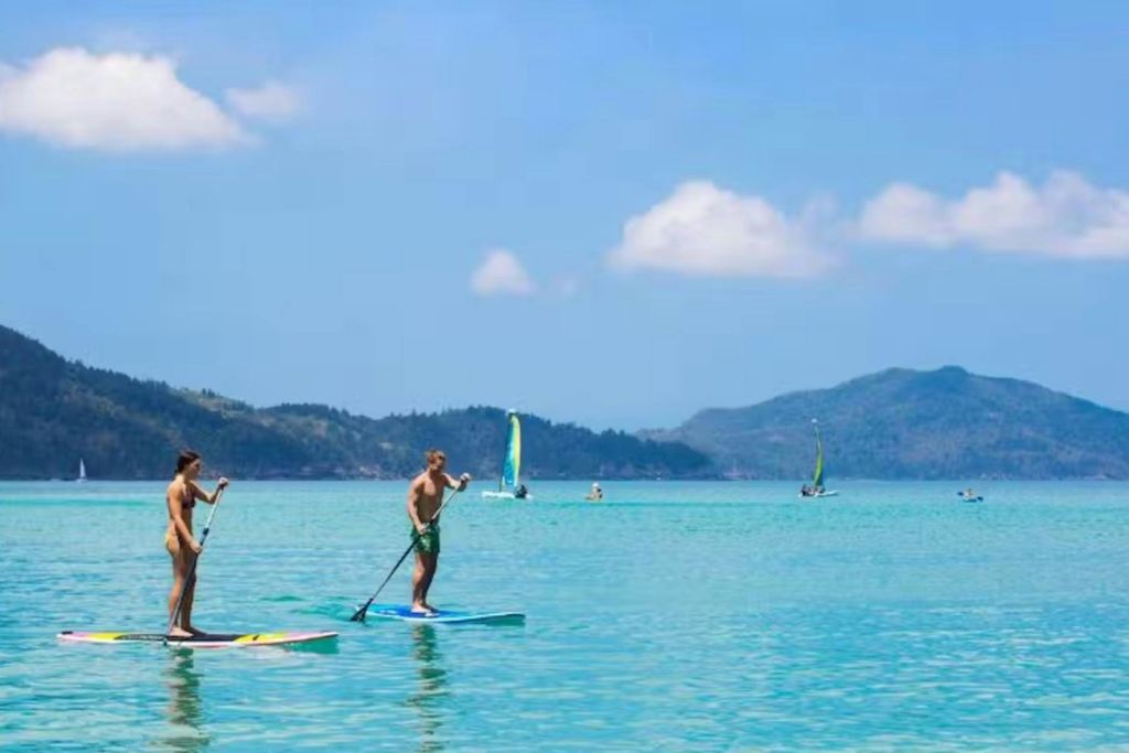 맑고 푸른 바다에서 패들보딩을 즐기는 두 사람, 멀리 떨어진 요트와 화창한 하늘 아래 푸른 언덕.