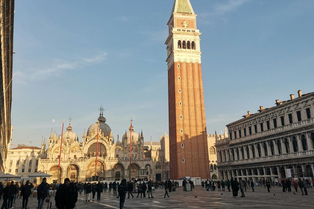 높이 솟은 시계탑과 화려한 대성당은 맑고 푸른 하늘 아래 분주한 공공 광장을 마주하고 있으며, 사람들이 걸어다니고 있습니다.