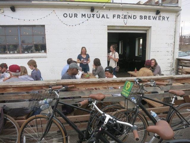 'Our Mutual Friend Brewery' 라고 적힌 흰색 건물 앞 야외 나무 테이블에 앉아 대화를 나눕니다. 인근에 자전거가 주차되어 있습니다.