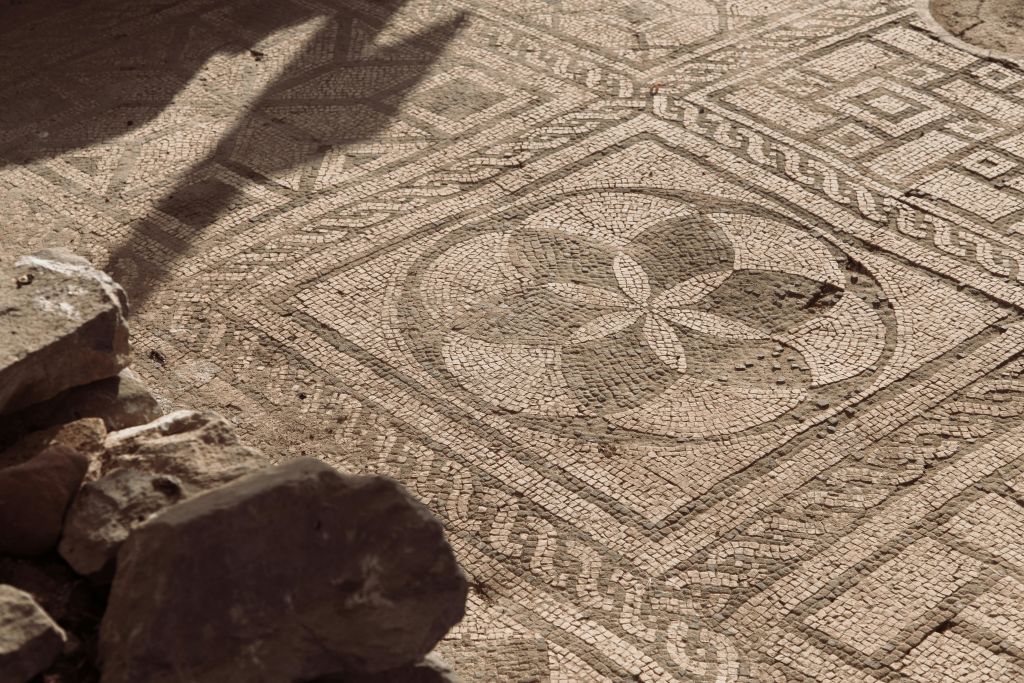 기하학적 패턴과 꽃무늬 디자인이 돋보이는 화려하고 오래된 모자이크 바닥은 부분적으로 머리 위 구조로 그늘져 있습니다.