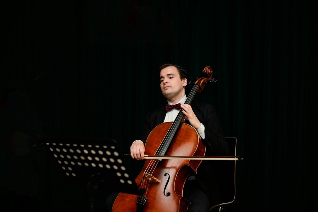 어두운 배경을 배경으로 정면에 음악 스탠드가 있는 정장 차림의 남성으로, 앉아서 첼로를 연주하고 있습니다.