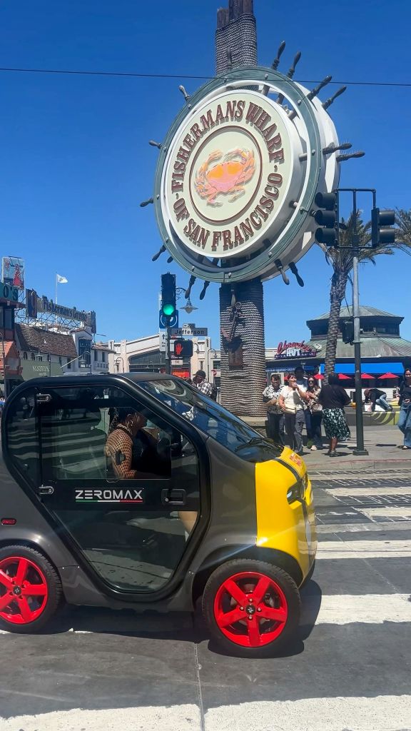 샌프란시스코의 피셔맨스 워프 (Fisherman's Wharf) 표지판을 지나가는 소형 전기 차량으로, 근처에 산책하는 사람들과 상점들이 있습니다.