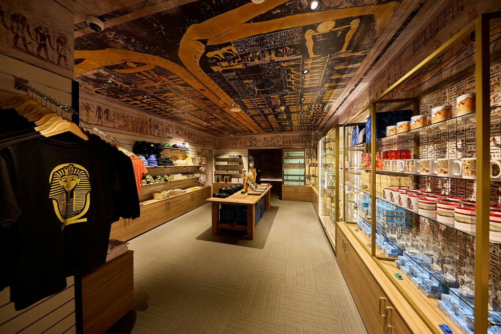 이집트 테마의 장식이 있는 선물 가게로, 옷이 진열된 선반, 기념품, 따뜻한 조명 아래 다양한 물품이 진열되어 있습니다.