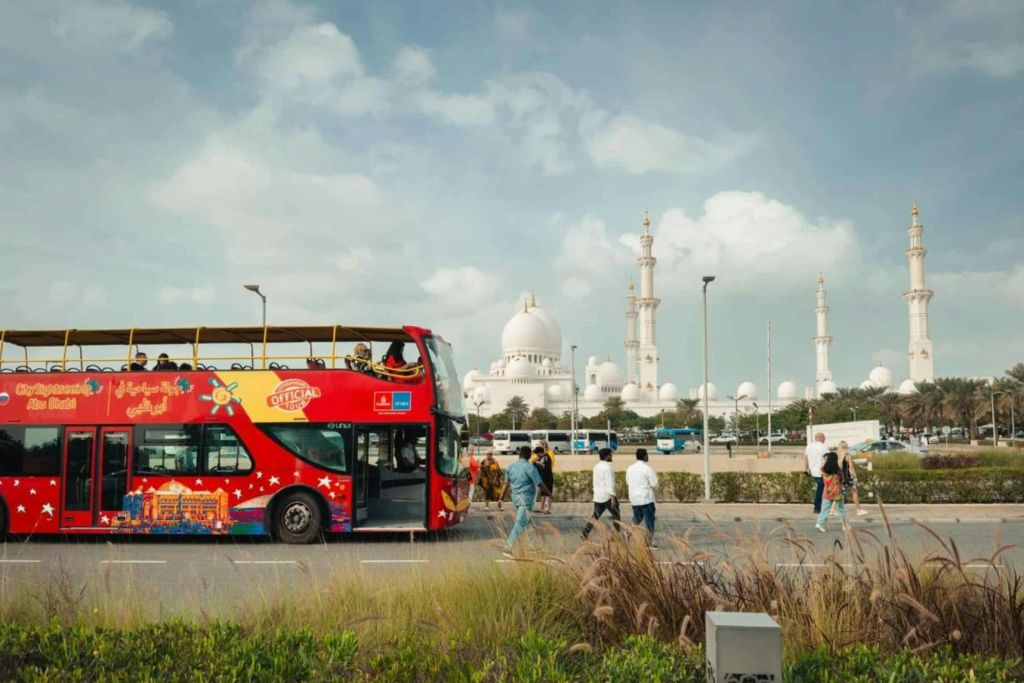 밝은 빨간색 2층 투어 버스, 승객, 근처 산책하는 사람들, 미나레트를 배경으로 한 커다란 흰색 모스크.