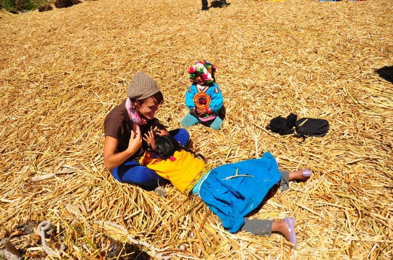 한 여성과 두 자녀가 짚으로 덮인 바닥에 앉아 있습니다. 한 여성과 한 자녀는 상호 작용하고 다른 한 자녀는 화려한 옷을 입고 있습니다.