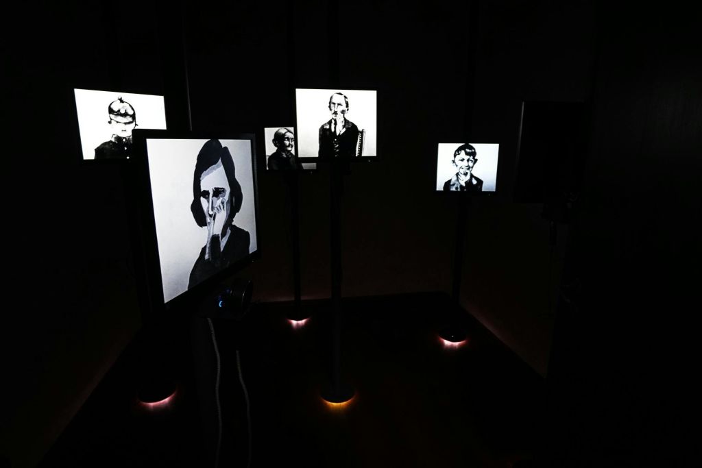 여러 개의 조명 스크린이 있는 어두운 방에는 사람들의 흑백 초상화가 전시되어 있습니다.