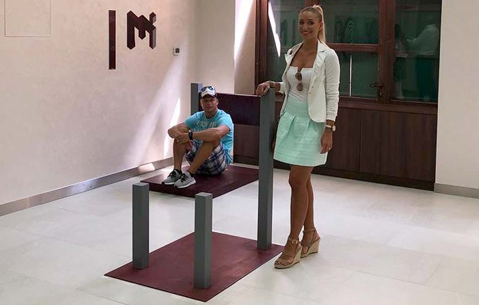 한 여성이 착시 현상 구조물 옆에 서서 미소 짓고 있으며, 한 남성이 플랫폼을 배경으로 앉아 있습니다.