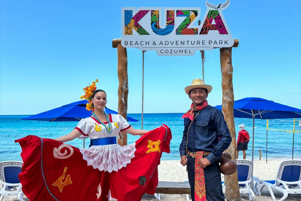 전통적인 드레스를 입은 여성과 모자를 쓴 남성이 "KUZU Beach & Adventure Cozumel" 표지판 아래 해변에서 포즈를 취하고 있습니다. 푸른색 의자와 바다를 배경으로.