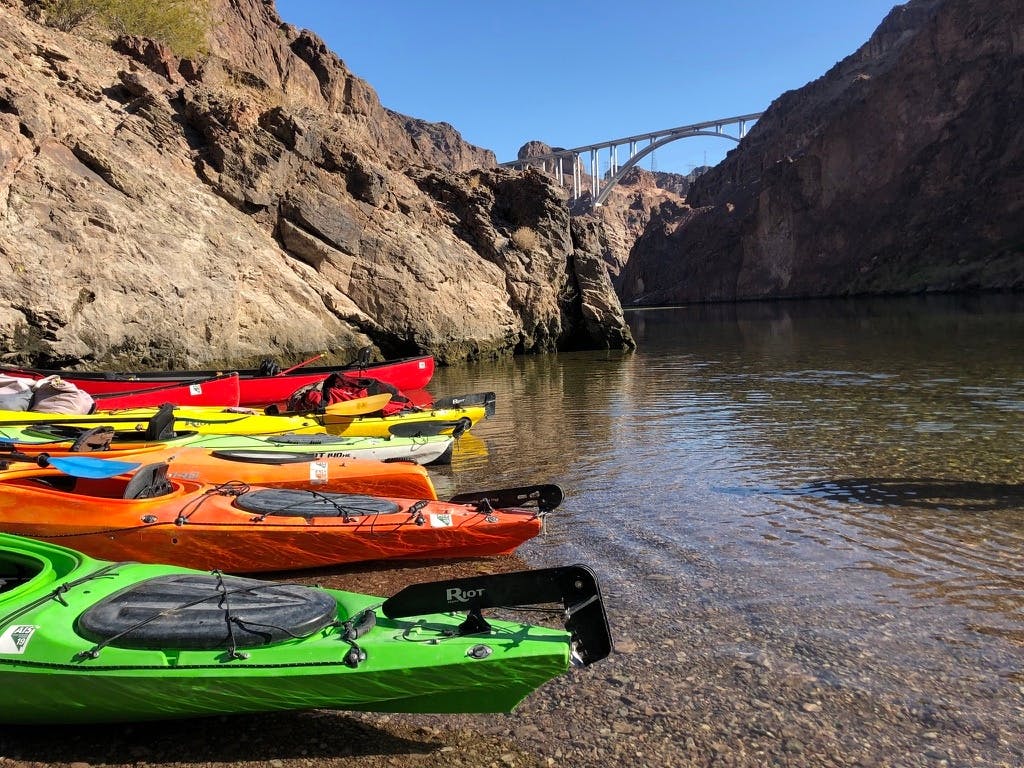 절벽으로 둘러싸인 잔잔한 물 근처의 바위 해안에 다채로운 색상의 카약이 줄지어 있고, 배경에는 다리가 보입니다.