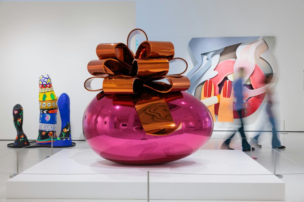 주황색 메탈릭 리본이 달린 선물을 형상화한 커다랗고 반짝이는 분홍색 조형물이 미술관에 전시되어 있습니다.