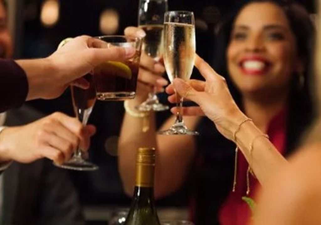 토스트에서 세 잔을 들어 올린 여성이 백그라운드에서 미소를 짓고 있으며, 앞쪽에 와인병이 부분적으로 보입니다.